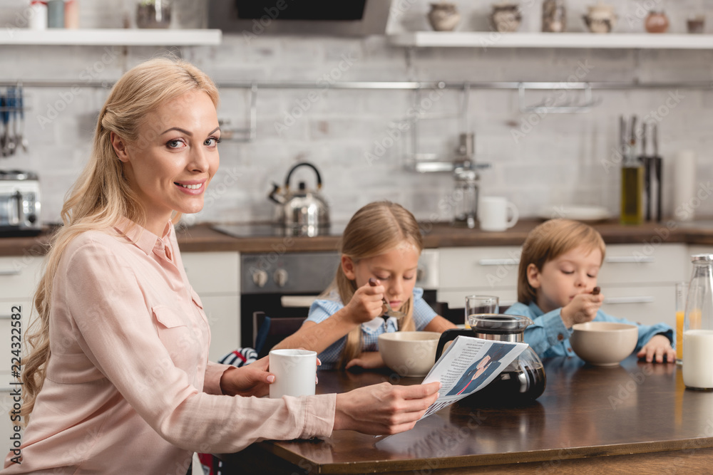 Obraz premium smiling mother having cup of coffee while sitting at kitchen with kids during breakfast and looking at camera