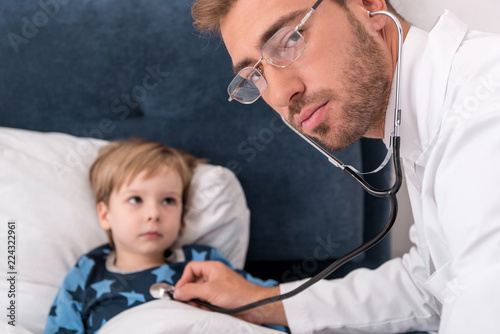 pediatrician litening childs breath with stethoscope and looking at camera while he lying in bed