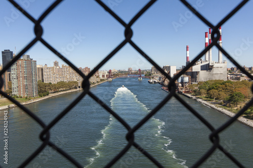 View from the Queensboro Bridge through a fence. looking over the East River with a boat passing by. Apartment buildings of Roosevelt Island and the Ravenswood Generating Station on Long Island City