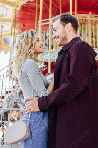 side view of affectionate couple in autumn outfit cuddling near carousel in amusement park
