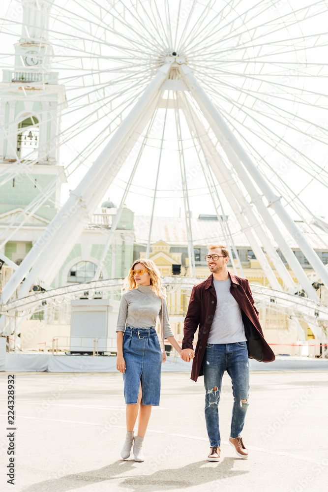 romantic couple in autumn outfit holding hands and walking near observation wheel