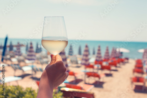 Female hand with a glass of white wine on the background of a beautiful beach. Travel vacations concept. Toned