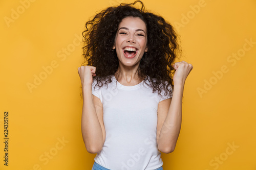 Image of young woman 20s with curly hair yelling and clenching fists, isolated over yellow background