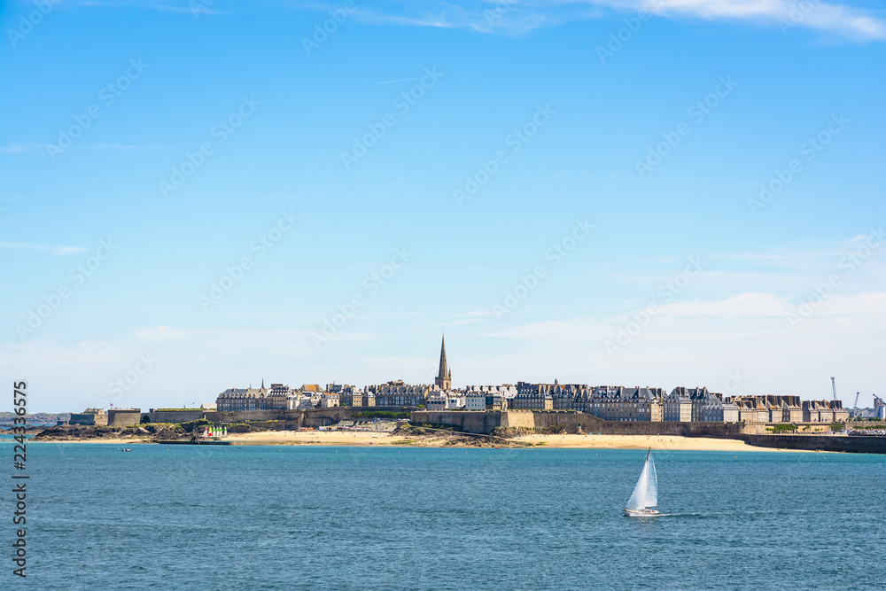 General view of the walled city of Saint-Malo in Brittany, France, with the steeple of the cathedral protruding above the buildings behind the wall and a boat sailing on the sea in the foreground.