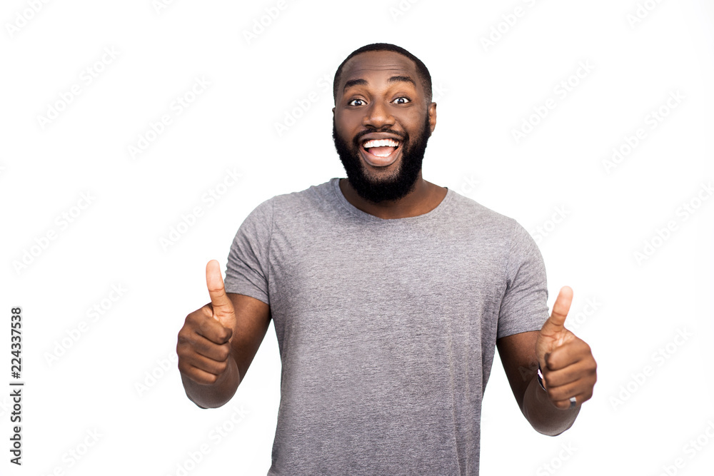 Indor shot of funny and emotive joyful dark-skinned guy with beard in trendy outfit, showing thumbs up and turning face at camera, smiling broadly, liking great and awesome party over white background Indor shot of funny and emotive joyful dark-skinned guy with beard in trendy outfit, showing thumbs up and turning face at camera, smiling broadly, liking great and awesome party over white background