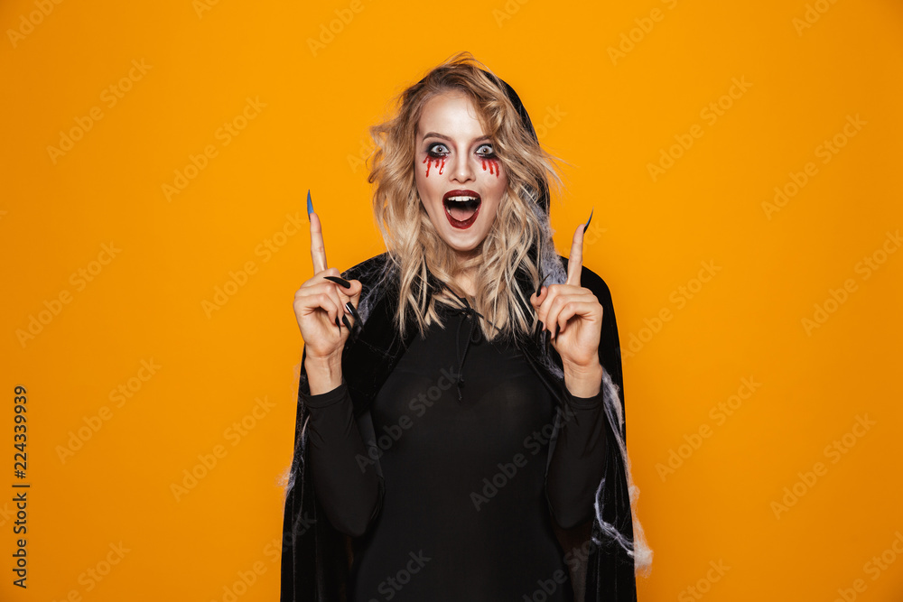 Obraz premium Amazing woman 20s wearing black costume and halloween makeup pointing fingers upward, isolated over yellow background