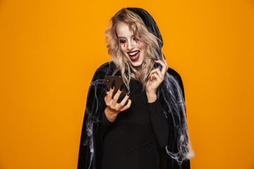 Scary wizard woman wearing black costume and halloween makeup holding smartphone, isolated over yellow background