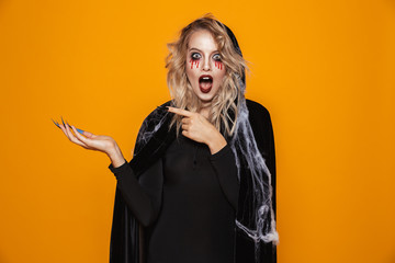 Surprised woman wearing black costume and halloween makeup holding copyspace on palm, isolated over yellow background