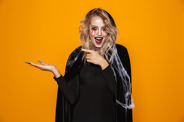 Frightening woman wearing black costume and halloween makeup holding copyspace on palm, isolated over yellow background