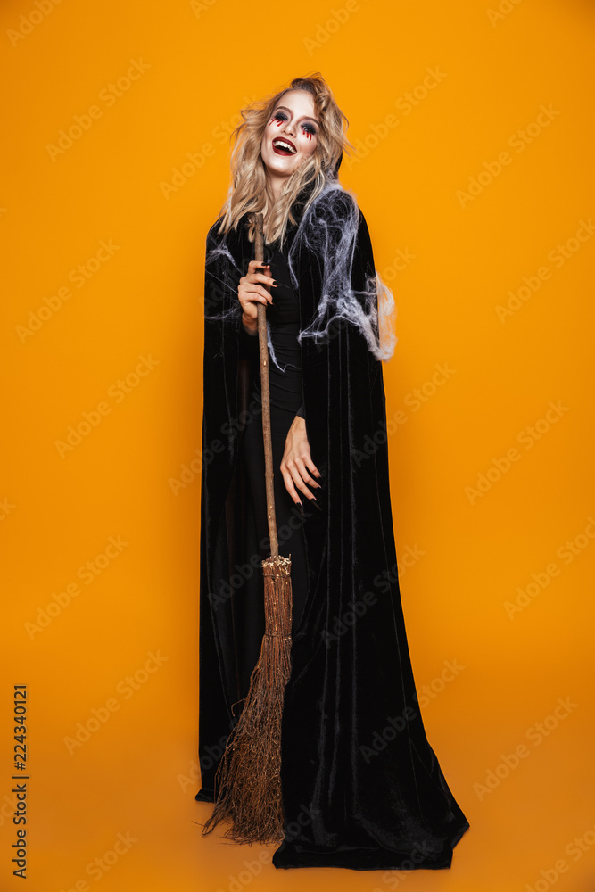 Full length image of dangerous witch woman wearing black costume and halloween makeup holding broom, isolated over yellow background Full length image of dangerous witch woman wearing black costume and halloween makeup holding broom, isolated over yellow background