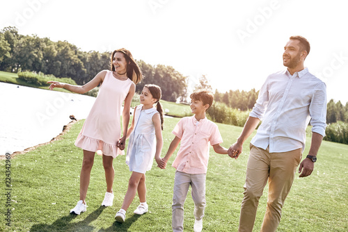 Bonding. Family of four walking on a grassy field near lake hold