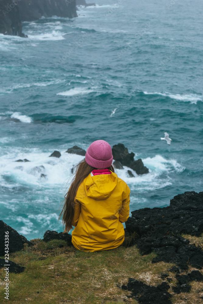 rear view of woman in yellow raincoat sitting on cliff in front of stormy ocean, iceland rear view of woman in yellow raincoat sitting on cliff in front of stormy ocean, iceland