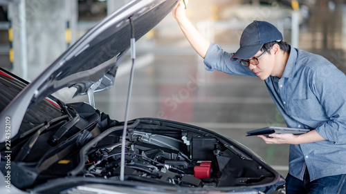 Asian auto mechanic holding digital tablet checking car engine under the hood in auto service garage. Mechanical maintenance engineer working in automotive industry. Automobile servicing and repair