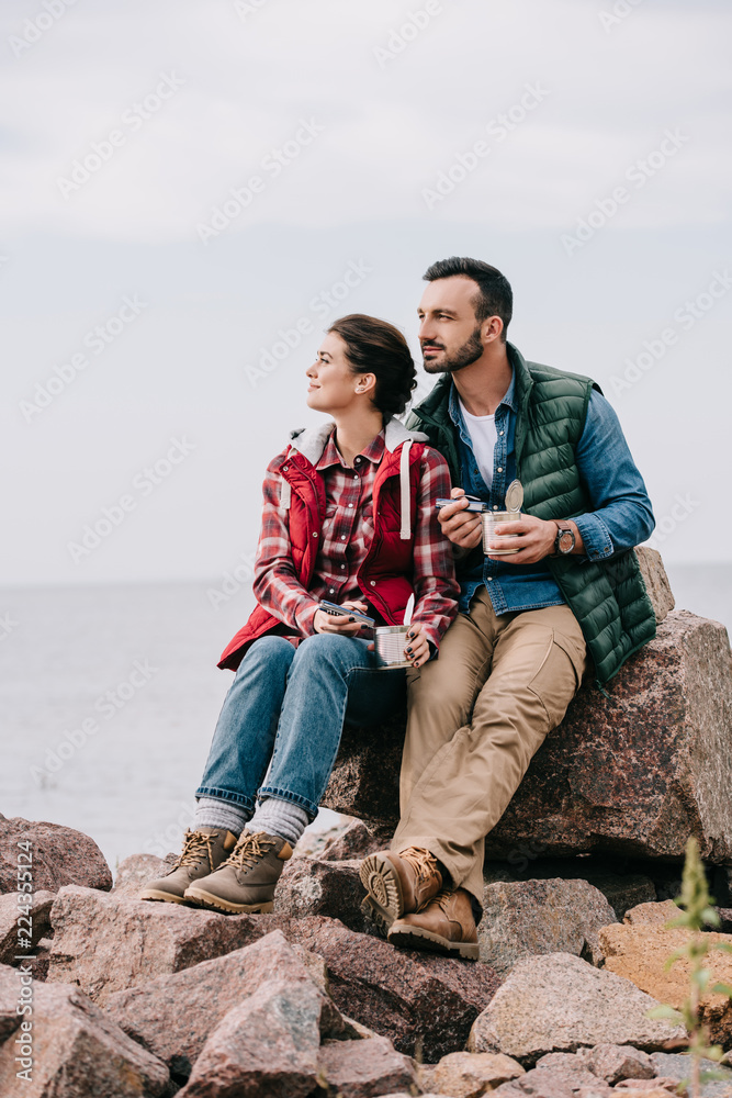 pensive couple of travelers with food in cans resting on rocks on sandy beach