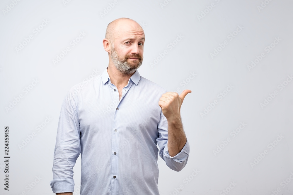 Handsome bearded man in casual clothes is pointing awaywith thumb looking at camera on gray background Handsome bearded man in casual clothes is pointing awaywith thumb looking at camera on gray background