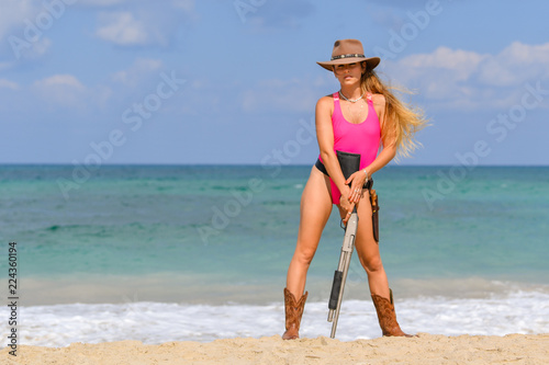 Beautiful sexy girl in a swimsuit with a carbine stands on the beach
