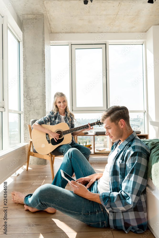 Fototapeta premium young man using digital tablet while girlfriend playing acoustic guitar at home