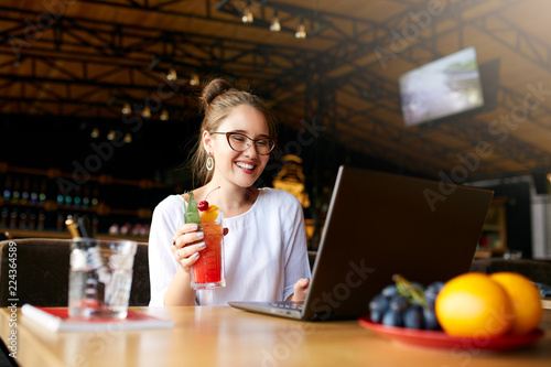 Smiling mixed race woman with cocktail in hand works with laptop. Businesswoman in glasses drinks juice for body hydration while working. Attractive designer quench thirst. Healthy lifestyle theme.