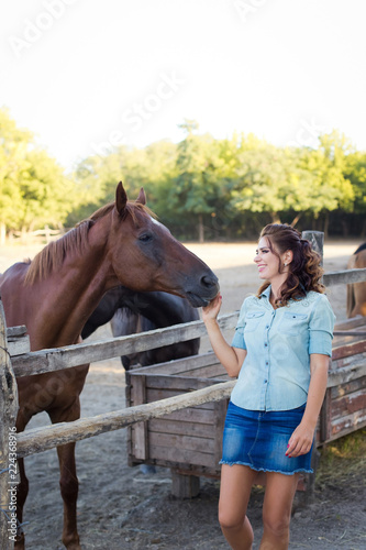 A young smiling woman with curly hair dressed in jeans at the stable with horses