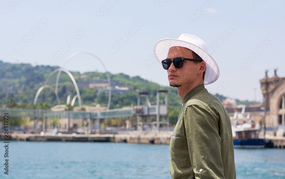 Young man in a hat, sunglasses stands in the port of Barcelona at the background of a monument to the waves and port customs, Barcelona