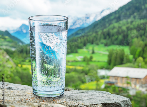 Glass of water on the stone. Blurred snow mountains tops and green forests at the background.