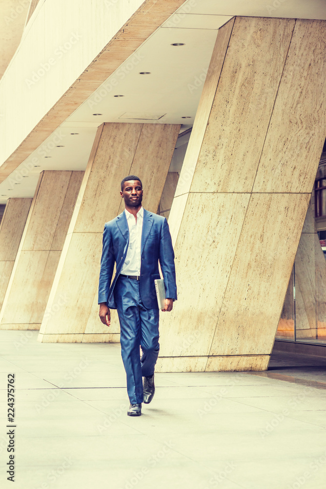 Young Handsome African American Businessman traveling, working in New York, wearing blue suit, white undershirt, leather shoes, holding laptop computer, walking on street outside office building..