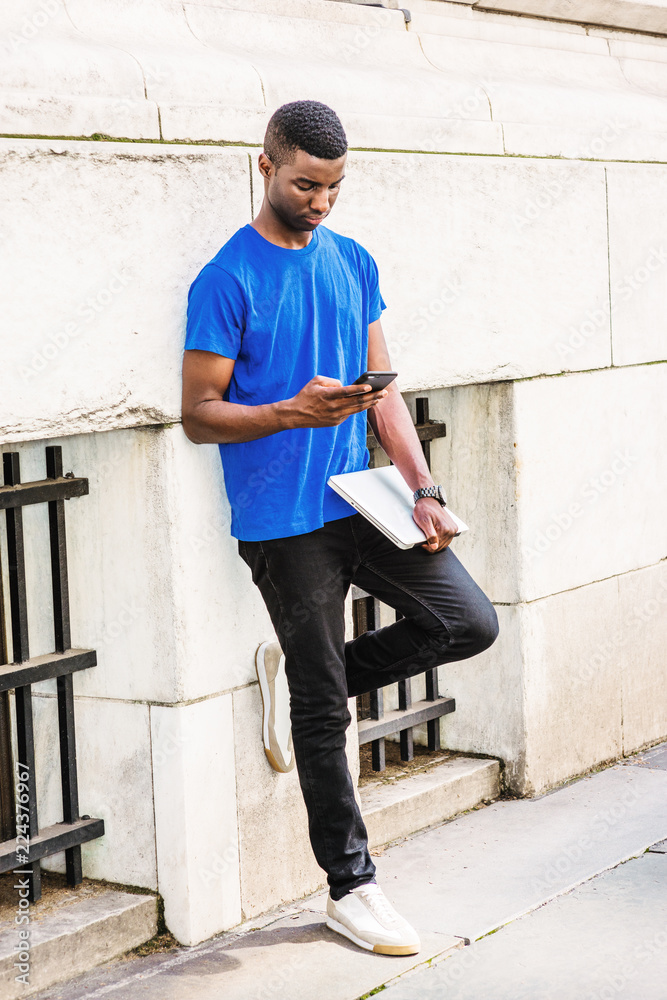 Young African American college student studying in New York, wearing blue T  suit, black pants, sneakers, holding laptop computer, standing outside  against wall of office building, texting.. Stock Photo | Adobe Stock