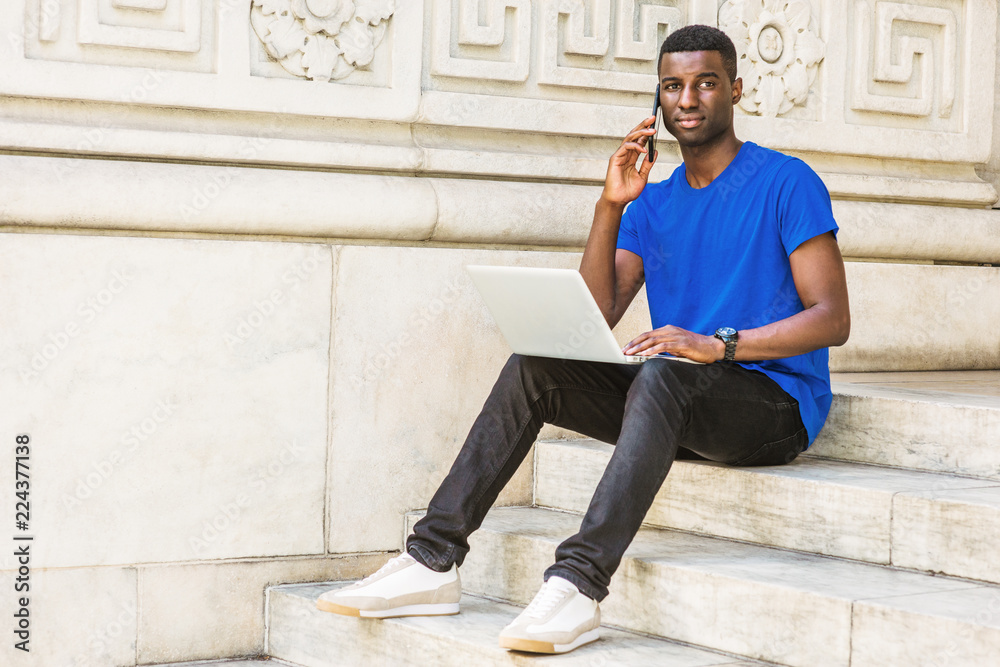 Young African American College Student studying in New York, wearing blue T shirt, black pants, sneakers, wristwatch, sitting on stairs on campus, working on laptop computer, talking on cell phone..