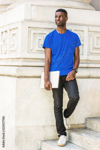 Obraz na płótnie Young African American Graduate Student studying in New York, wearing blue T shirt, black pants, sneakers, wristwatch, holding laptop computer, standing on stairs outside office building on campus.. w salonie