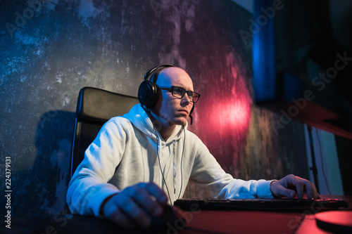 Portrait of concentrated young man in glasses and headset in dark room playing online computer game or competing in online esport tournament