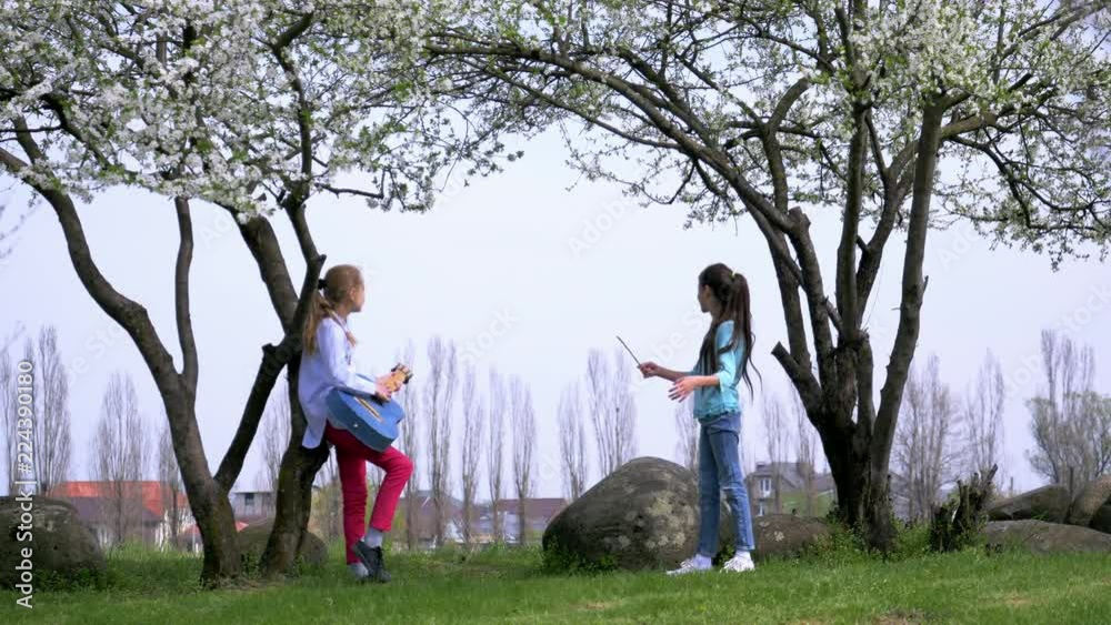two funny little girlfriends play guitar and orchestrate conduct a stick outdoors. beautiful view springtime flowering tree in countryside two funny little girlfriends play guitar and orchestrate conduct a stick outdoors. beautiful view springtime flowering tree in countryside