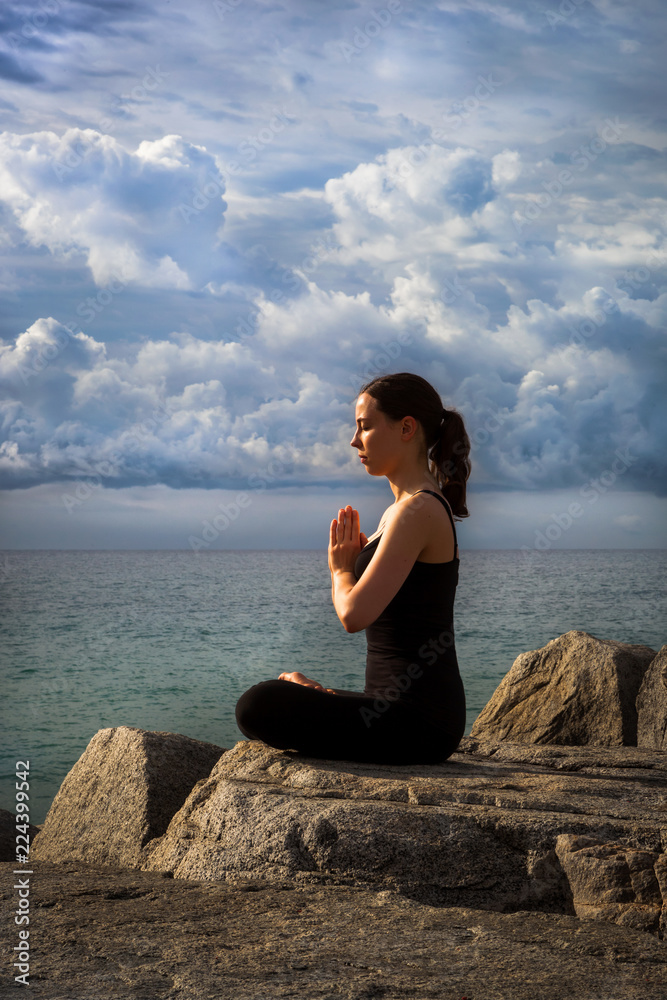 Young woman practicing yoga on rocks at sunrise with dramatic sky