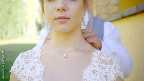 groom is putting golden chain with white pearl on neck of his young bride and fastening, woman is looking photo