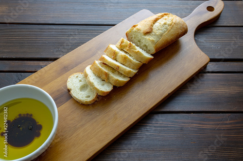 A french bread baguette sliced up on a cutting board with a bowl of olive oil with a splash of balsamic vinegar along side a bottle of olive oil that had a stem of rosemary.