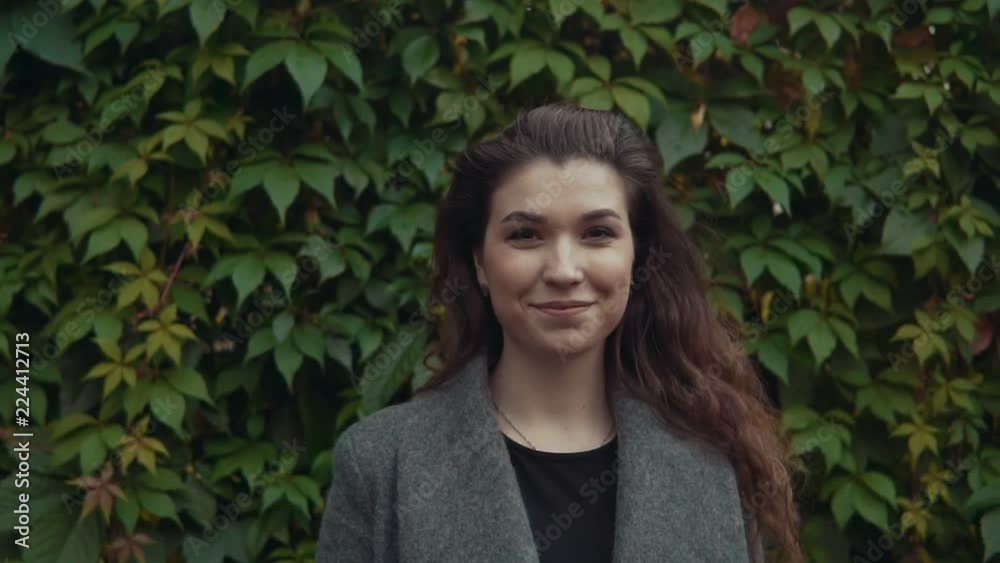 Young beautiful brunette woman in coat smiling and looking at camera in slow motion at background of green leaves. Close up shot Young beautiful brunette woman in coat smiling and looking at camera in slow motion at background of green leaves. Close up shot