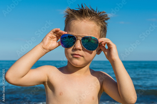 a beautiful little boy posing on a beach by the sea with sunglasses