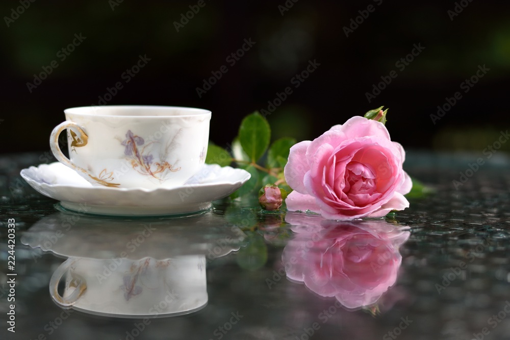 On the glass table, wet from rain, rose flower and antique cup of tea.