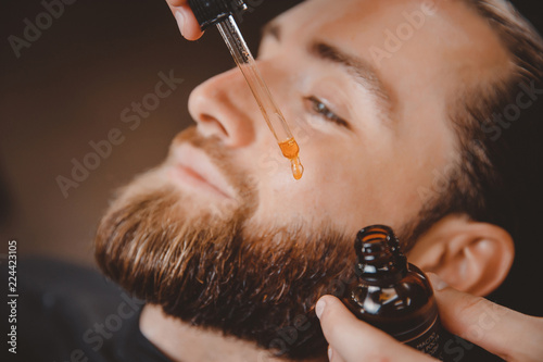 Fotografie Barber applies beard oil with dropper for man in barbarshop.