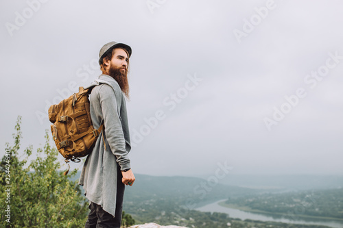 Young Tourist stands on a cliff face and looks at nature.Bearded man wore a cool hat and a stylish backpack