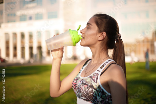 Beautiful brunette sports fitness slim woman in top with tropical print drinking water outdoors in the park on green grass with sundown light.