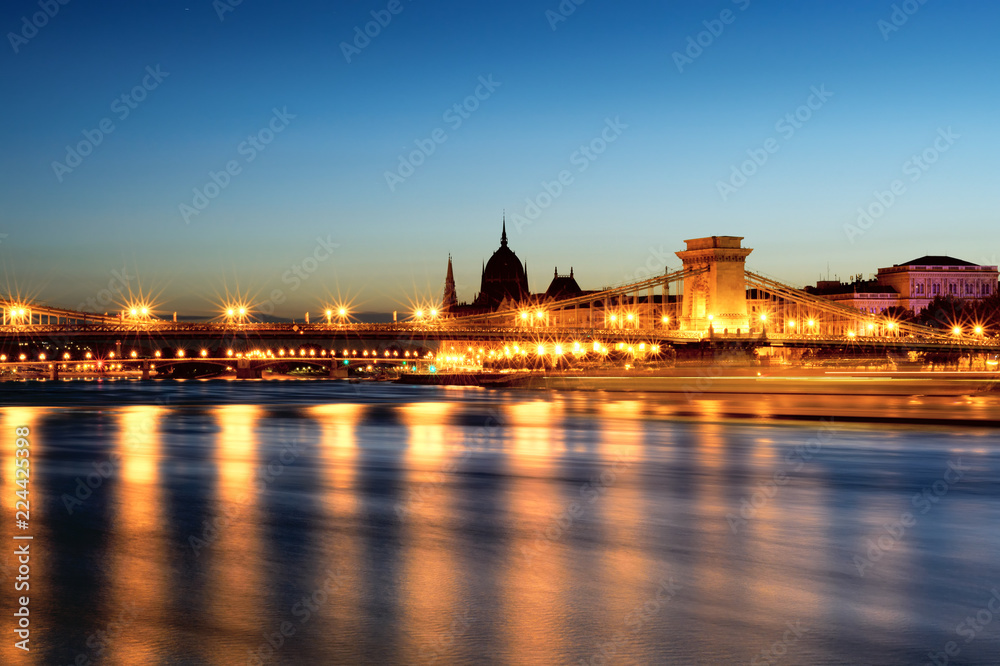 Illuminated Chain bridge reflecting in Danube river, silhouette of Parliament domes