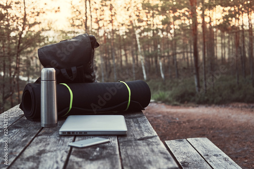 Travel concept. Backpack, tourist mat, laptop, smartphone and thermos bottle on a wooden table in beautiful forest.
