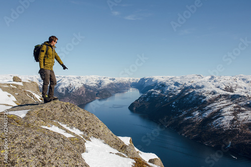 Young hiker man standing on the big rock and pointing on the fjord and mountains. Preikestolen, Pulpit rock, Lysefjord, Norway. Sunny winter day