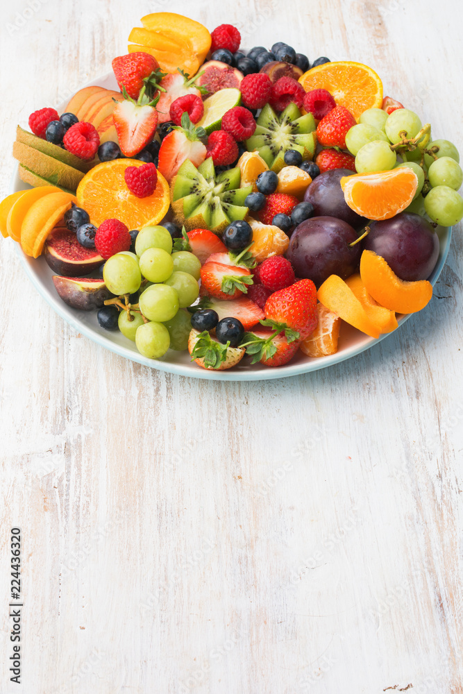 Healthy fruit platter, strawberries raspberries oranges plums apples kiwis grapes blueberries on the white wooden table, vertical, copy space for text, selective focus