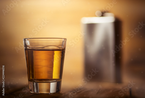 Whiskey shot glass on top of a wooden table background and a metal hip flask in the background