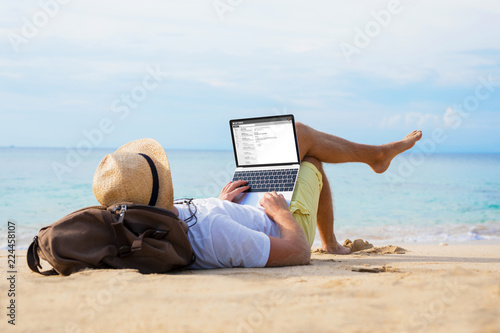 Man reading email on laptop while relaxing on beach