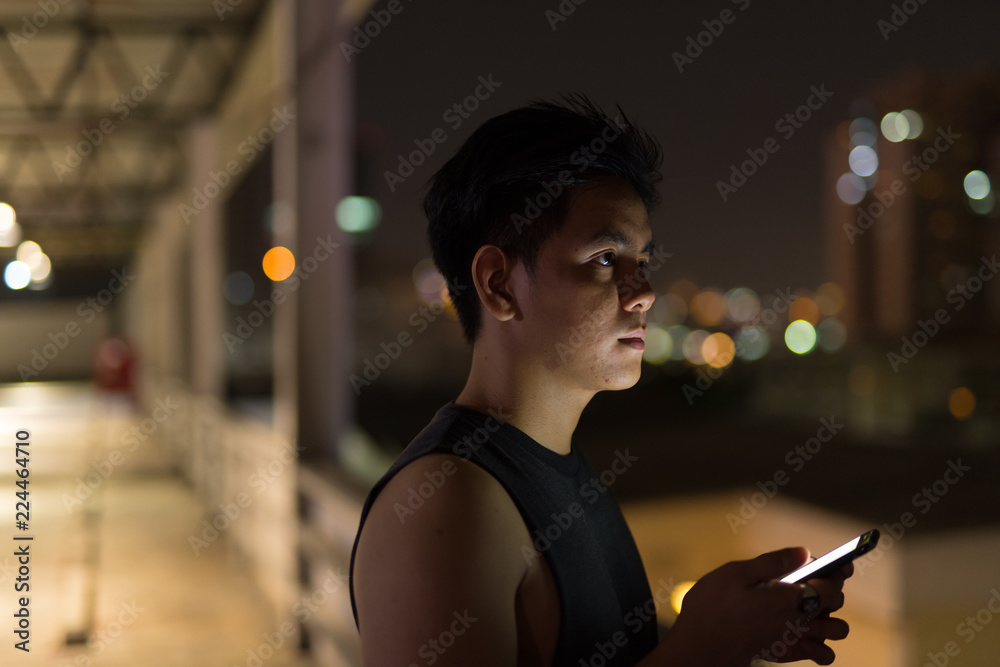 Fototapeta premium Young handsome Asian man thinking while using phone against view of the city at night