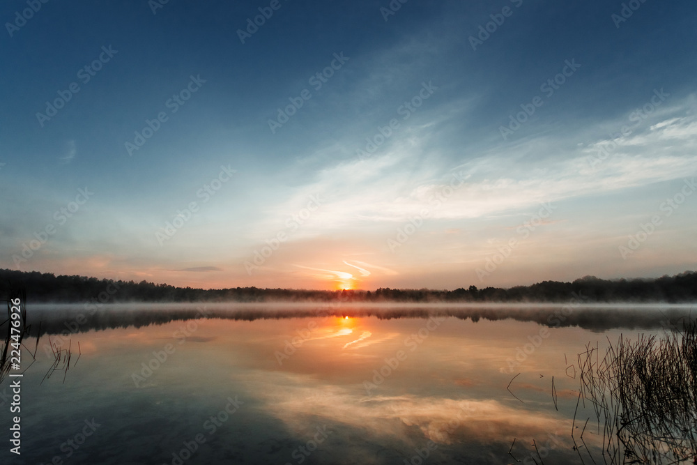 Beautiful, red dawn on the lake. The rays of the sun through the fog. The blue sky over the lake, the morning comes, the sky is reflected in the water.