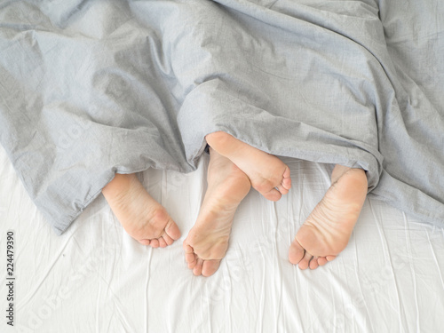 Feet of couple sleeping side by side in comfortable bed. Close up of feet in a bed under white blanket. Bare feet of a man and a woman peeking out from under the cove.