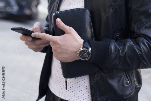 young man wearing leather jacket and wrist watches holding a leather wallet and using a smartphone 